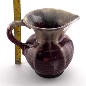 May include: A vintage ceramic pitcher with a handle and a spout. The pitcher is primarily burgundy with cream and gray accents. The handle has a textured design. The pitcher is approximately 5 inches tall.