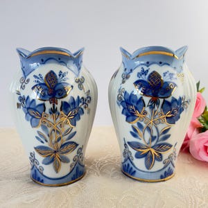 May include: A pair of white ceramic vases featuring blue floral patterns and gold detailing. The vases have a flared top with a blue rim and a gold band. The floral design includes blue flowers and gold leaves.