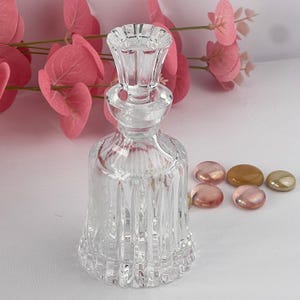 May include: A clear glass bottle with a stopper, featuring vertical ribbed detailing. Several small, iridescent, and gold-toned glass pebbles are next to the bottle. Pink foliage is in the background.