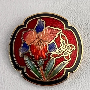 May include: A round, enamelled brooch featuring a floral design with a butterfly. The brooch has a red background with a black border and gold trim. The flower has orange, blue, and red petals with green leaves.