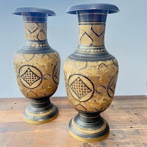May include: Two decorative vases in black and gold. The vases have a flared top and a wide base. The gold sections feature an intricate leaf and geometric pattern. The vases are made of metal and are suitable for home decor.