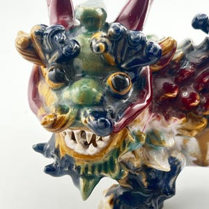 May include: A detailed ceramic dragon figurine with a colourful glaze. The dragon features a green face, dark blue accents, and red horns. Its mouth is open, revealing white teeth. The figurine is a decorative item.