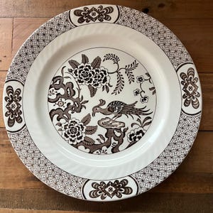 May include: A white ceramic dinner plate with a brown and black floral design featuring a bird. The plate has a decorative border with a repeating geometric pattern.