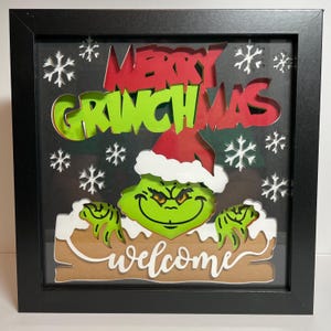 May include: A framed 3D papercraft of the Grinch with a red Santa hat, peeking over a sign that says "Welcome" and a banner that says "Merry Grinchmas". The background is black with white snowflakes.