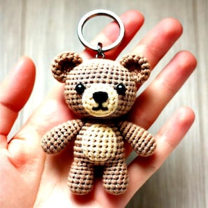 Op de afbeelding: Een kleine bruine gehaakte teddybeer sleutelhanger met zwarte ogen en een zwarte neus. De beer houdt een zilveren sleutelring vast.