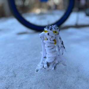 Puede incluir: Una criatura blanca de limpiapipas con ojos negros y detalles amarillos se encuentra en la nieve. La criatura está hecha de limpiapipas y tiene un cuerpo largo y delgado. La criatura está de pie en la nieve, que es blanca y esponjosa.