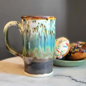 Puede incluir: Una taza de cerámica alta con un esmalte único en tonos marrón, blanco, azul y verde. La taza tiene un asa grande y una base blanca. Dos donuts con chispas de colores están en un plato verde pequeño.