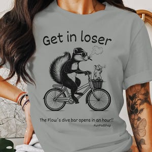 May include: Gray t-shirt with a black and white illustration of a skunk riding a bicycle with a rat passenger. The text "Get in loser" and "The Flow's dive bar opens in an hour!" is printed on the shirt.