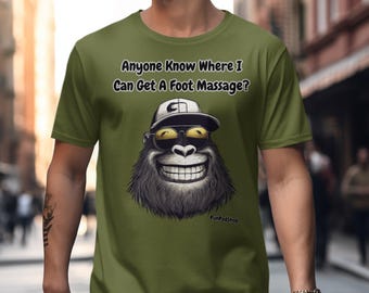 May include: A green t-shirt with a cartoon gorilla wearing sunglasses and a baseball cap. The text on the shirt reads "Anyone know where I can get a foot massage?"