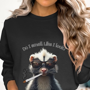 May include: Black sweatshirt with a white graphic of a skunk wearing sunglasses and smoking a cigarette. The text "Do I smell like I look?" is printed on the sweatshirt.