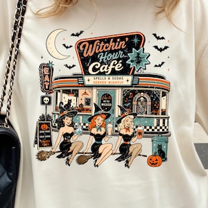 Pode incluir: Uma t-shirt em tom creme com uma estampa gráfica de um restaurante chamado Witchin' Hour Cafe, três bruxas, uma lua crescente, morcegos e uma abóbora. A peça é combinada com uma bolsa de ombro preta acolchoada com corrente dourada.