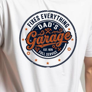 Könnte beinhalten: Weißes T-Shirt mit einem kreisförmigen Aufdruck „Dad's Garage“ in Orange und Blau. Der Aufdruck enthält den Text „Fixes Everything“ und „Full Service“ in einem seilartigen Rahmen, mit dem Gründungsjahr „Est. 1978“.