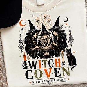 Puede incluir: Una camiseta color crema con un gr&aacute;fico de tres brujas con sombreros negros reunidas alrededor de un caldero. El dise&ntilde;o incluye calaveras, estrellas, lunas, velas y un gato negro. El texto dice WITCH COVEN y MIDNIGHT RITUAL SOCIETY.