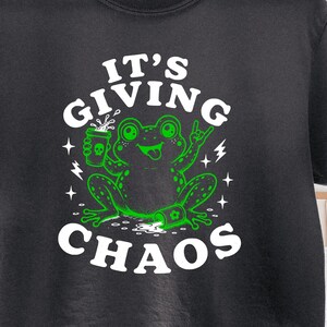 Op de afbeelding: Donkergrijs t-shirt met een groene kikker afbeelding en de tekst "IT'S GIVING CHAOS". De kikker houdt een drankje vast en heeft een vredesteken. Het ontwerp bevat bliksemschichten en sterren.