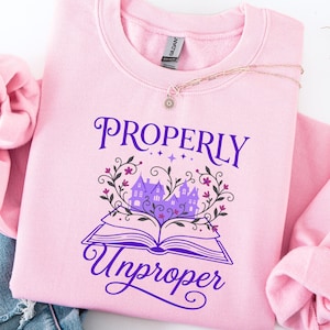 May include: A light pink sweatshirt with the words "Properly Unproper" in purple script. The design features an open book with a castle and floral accents. A silver necklace with a pendant rests on the sweatshirt.