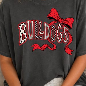 May include: A charcoal gray short-sleeve t-shirt featuring the word BULLDOGS in a bold, black-and-white polka dot and striped pattern. The text is accented with a large, bright red bow with black polka dots that wraps around the lettering.