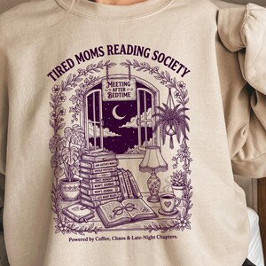 Op de afbeelding: Beige sweatshirt met een paarse afbeelding. De afbeelding toont een venster met een maan, boeken en planten. Tekst bevat "TIRED MOMS READING SOCIETY" en "MEETING AFTER BEDTIME". De zin "Powered by Coffee, Chaos & Late-Night Chapters" is ook aanwezig.
