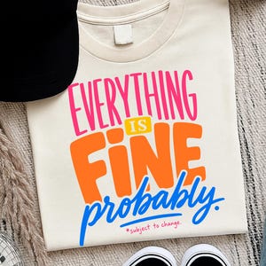May include: A cream-colored t-shirt featuring the text Everything IS Fine probably in bright pink, orange, and blue lettering, with the smaller text *subject to change. printed in red at the bottom of the graphic design on the front of the shirt.