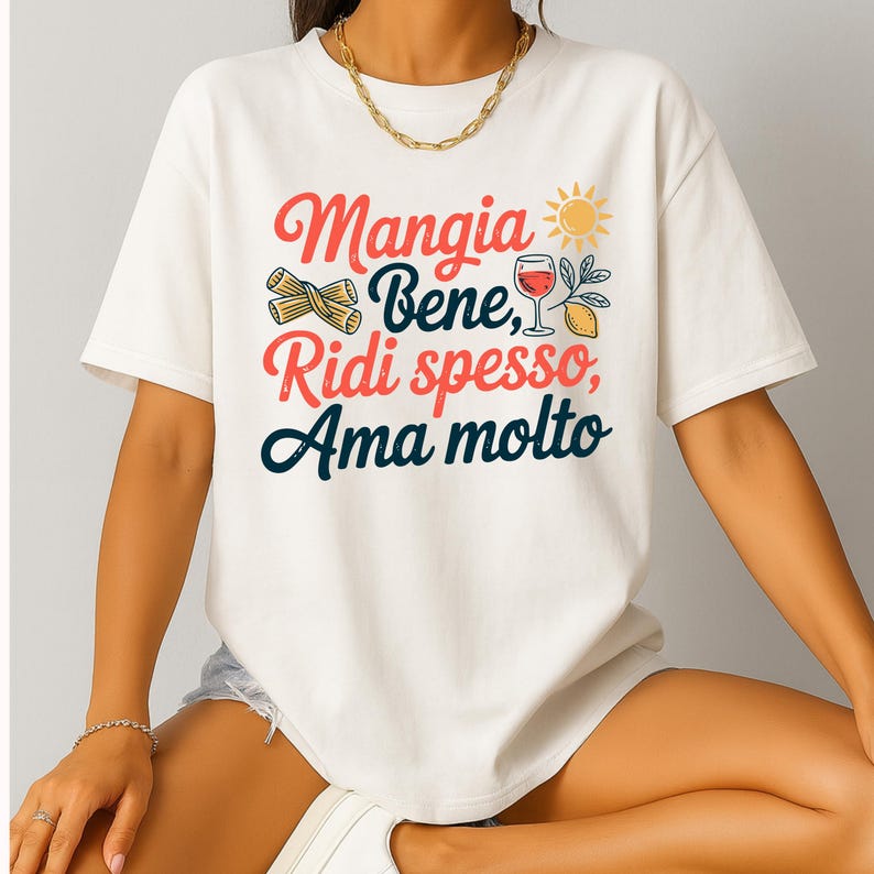 May include: White t-shirt with the Italian phrase "Mangia Bene, Ridi spesso, Ama molto" in red, blue, and green. The design includes pasta, a glass of wine, a lemon, and a sun. The shirt is a casual, everyday wear item.
