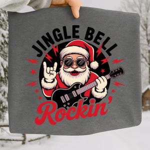 Puede incluir: Bolso tote gris con temática navideña que presenta un gráfico de Papá Noel con gafas de sol tocando una guitarra eléctrica negra. El texto "JINGLE BELL Rockin'" está impreso encima y debajo de la ilustración de Papá Noel.