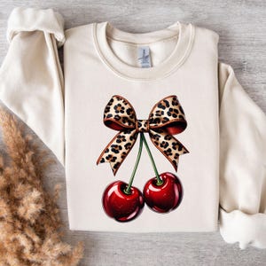 Puede incluir: Una sudadera de color crema con un lazo de estampado de leopardo y dos cerezas rojas. El lazo está atado alrededor de los tallos de las cerezas.