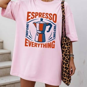 May include: A light pink t-shirt with the words "ESPRESSO BEFORE EVERYTHING" in red and blue. The graphic features a coffee maker illustration. A leopard print tote bag hangs on the right side.