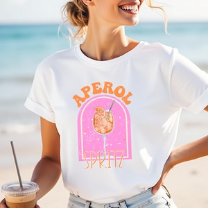 Puede incluir: Camiseta blanca con un gráfico retro de un cóctel Aperol Spritz. El diseño presenta las palabras "APEROL" y "SPRITZ" en naranja y blanco, con un arco rosa y una ilustración de cóctel. La camiseta se lleva en la playa.