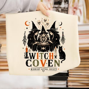 Puede incluir: Un art&iacute;culo de tela color crema con un gr&aacute;fico negro y naranja de tres brujas con sombreros puntiagudos alrededor de un caldero. El dise&ntilde;o incluye calaveras, estrellas, lunas, velas, un gato negro y el texto WITCH COVEN MIDNIGHT RITUAL SOCIETY.