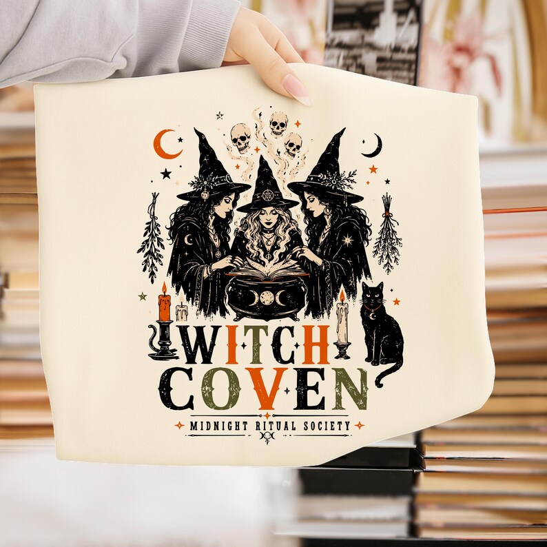 Puede incluir: Un art&iacute;culo de tela color crema con un gr&aacute;fico negro y naranja de tres brujas con sombreros puntiagudos alrededor de un caldero. El dise&ntilde;o incluye calaveras, estrellas, lunas, velas, un gato negro y el texto WITCH COVEN MIDNIGHT RITUAL SOCIETY.