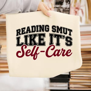 May include: Cream-colored tote bag with the text "READING SMUT LIKE IT'S Self-Care" in black and burgundy lettering. The bag is held up by a person. The text is in a bold, collegiate-style font.