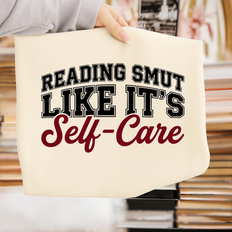 May include: Cream-colored tote bag with the text "READING SMUT LIKE IT'S Self-Care" in black and burgundy lettering. The bag is held up by a person. The text is in a bold, collegiate-style font.
