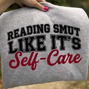 May include: A heather gray sweatshirt with the text "READING SMUT LIKE IT'S Self-Care" in black and burgundy lettering. The sweatshirt is held up by a hand with pink nail polish.