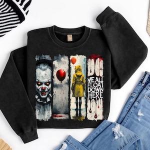 Può includere: Felpa nera con un design grafico raffigurante un clown, una persona con un impermeabile giallo e la scritta "WE ALL FLOAT DOWN HERE". Il design include palloncini rossi e dettagli di sangue che gocciola.