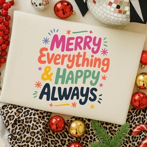 May include: A cream-colored pillow with the words "MERRY Everything & HAPPY ALWAYS" in colorful, playful lettering. The pillow is surrounded by Christmas ornaments, including red and gold baubles, and a leopard print fabric.