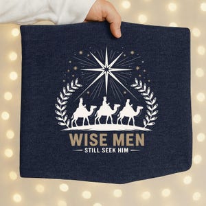 Puede incluir: Tela azul marino con un gráfico blanco de tres reyes magos sobre camellos bajo una estrella. El texto dice "WISE MEN - STILL SEEK HIM" en una fuente serif, rodeado por una corona de laurel.