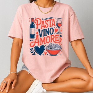 May include: A light pink t-shirt with the words "Pasta Vino Amore" in red and blue script. The design includes a wine bottle, a glass of wine, a bowl of pasta, and a blue heart.