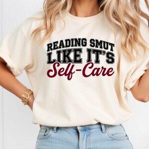 May include: Cream-colored t-shirt with the text "READING SMUT LIKE IT'S Self-Care" in black and burgundy lettering. The shirt has short sleeves and a crew neck. The overall style is casual and comfortable.