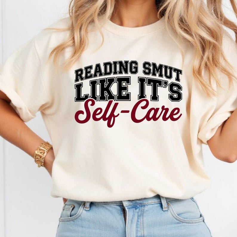 May include: Cream-colored t-shirt with the text "READING SMUT LIKE IT'S Self-Care" in black and burgundy lettering. The shirt has short sleeves and a crew neck. The overall style is casual and comfortable.