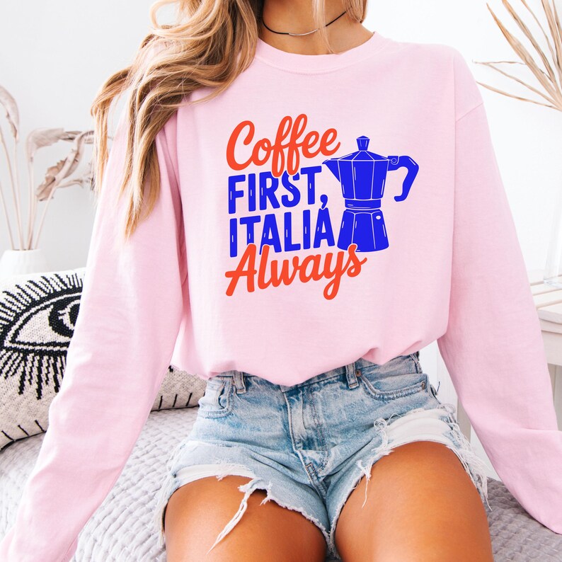 May include: A light pink sweatshirt with the text "Coffee FIRST, ITALIA Always" in red and blue. A blue coffee pot graphic is next to the text. The sweatshirt is long-sleeved.