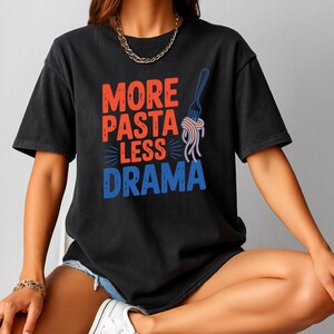 May include: Black t-shirt with the text "MORE PASTA LESS DRAMA" in red and blue. A blue fork with pasta strands is to the right of the text. The shirt is worn by a person.