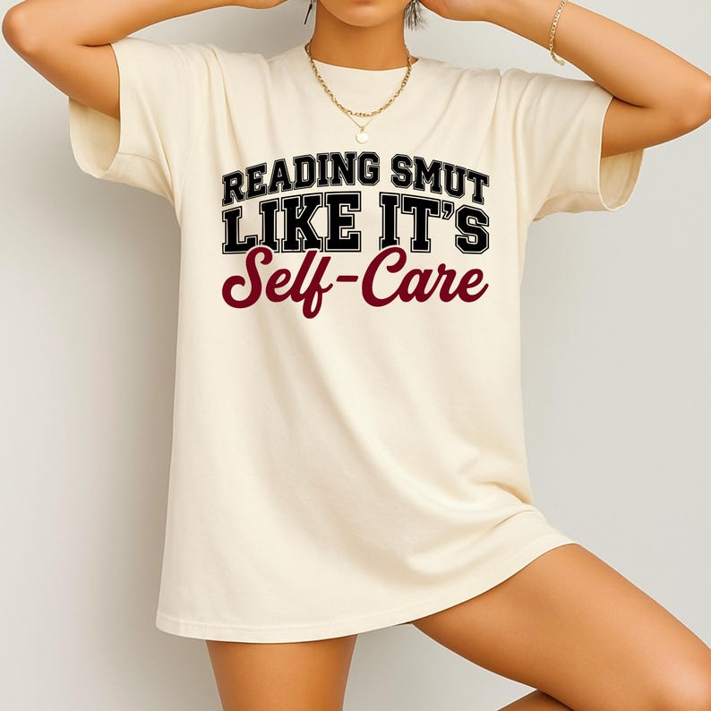 May include: Cream-colored t-shirt with the text "READING SMUT LIKE IT'S Self-Care" in black and burgundy lettering. The shirt has short sleeves and a relaxed fit.