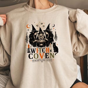 Puede incluir: Una sudadera beige de cuello redondo con un estampado gr&aacute;fico de tres brujas con sombreros puntiagudos reunidas alrededor de un caldero y un libro abierto. El dise&ntilde;o incluye calaveras, velas, un gato negro y el texto WITCH COVEN MIDNIGHT RITUAL SOCIETY.