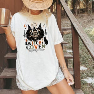 Puede incluir: Una camiseta de color blanco roto con un estampado gr&aacute;fico de tres brujas con sombreros puntiagudos reunidas alrededor de un caldero. El dise&ntilde;o incluye calaveras, un gato negro, velas y el texto "WITCH COVEN" con "MIDNIGHT RITUAL SOCIETY".