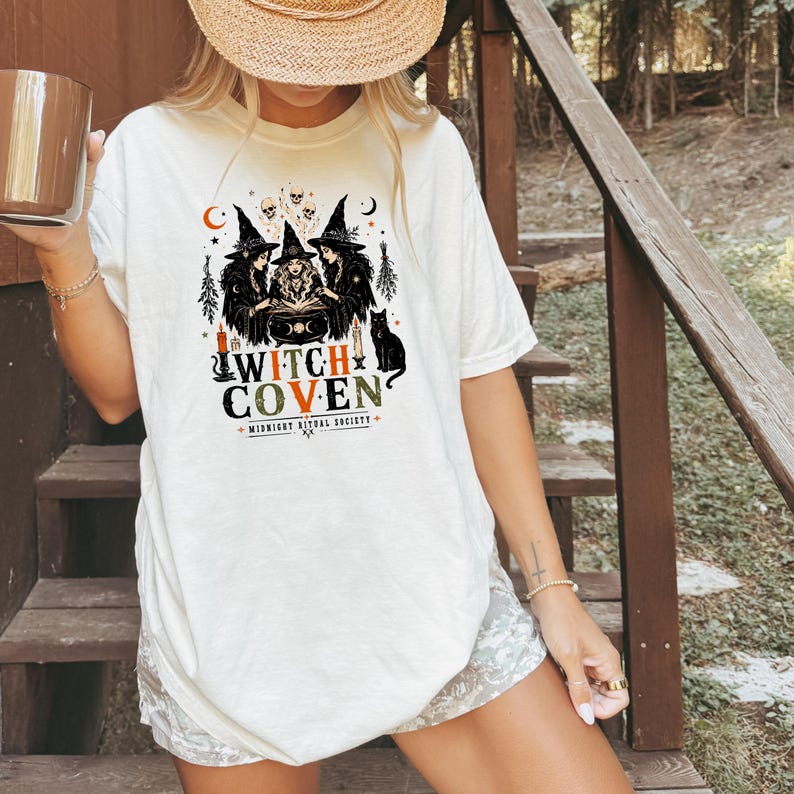 Puede incluir: Una camiseta de color blanco roto con un estampado gr&aacute;fico de tres brujas con sombreros puntiagudos reunidas alrededor de un caldero. El dise&ntilde;o incluye calaveras, un gato negro, velas y el texto "WITCH COVEN" con "MIDNIGHT RITUAL SOCIETY".