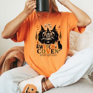 Puede incluir: Una camiseta naranja con un estampado gr&aacute;fico de tres brujas con sombreros puntiagudos alrededor de un caldero, con un gato negro, velas, calaveras y lunas. El texto dice WITCH COVEN y MIDNIGHT RITUAL SOCIETY en una fuente decorativa.