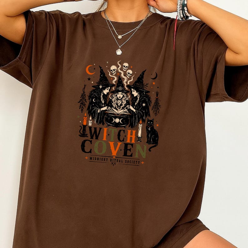Puede incluir: Una camiseta marr&oacute;n con un estampado gr&aacute;fico de tres brujas con sombreros puntiagudos reunidas alrededor de un caldero. El dise&ntilde;o incluye calaveras, un gato negro, velas y el texto "WITCH COVEN" con "MIDNIGHT RITUAL SOCIETY" debajo.