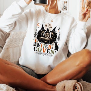 Puede incluir: Una sudadera blanca de manga larga con un estampado gr&aacute;fico de tres brujas alrededor de un caldero con calaveras, velas y un gato negro. El texto dice WITCH COVEN y MIDNIGHT RITUAL SOCIETY en letras de color negro, naranja y verde.