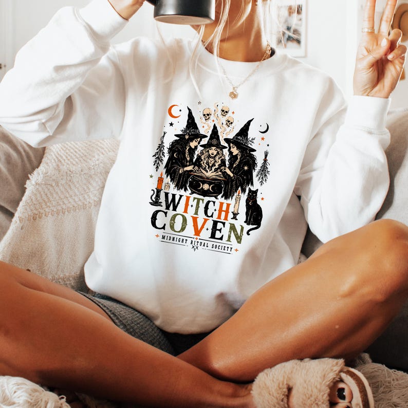Puede incluir: Una sudadera blanca de manga larga con un estampado gr&aacute;fico de tres brujas alrededor de un caldero con calaveras, velas y un gato negro. El texto dice WITCH COVEN y MIDNIGHT RITUAL SOCIETY en letras de color negro, naranja y verde.