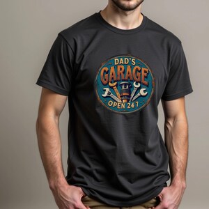Może przedstawiać: Czarny T-shirt z vintage'owym znakiem, na kt&oacute;rym widnieje napis "Dad's Garage Open 24/7". Znak ma niebieski tło z pomarańczowym tekstem i wzorem klucza.