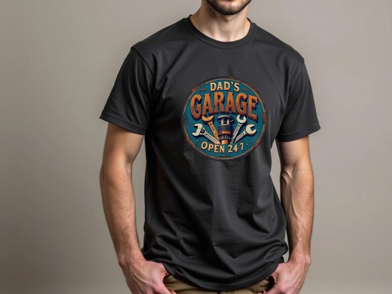 Może przedstawiać: Czarny T-shirt z vintage'owym znakiem, na kt&oacute;rym widnieje napis "Dad's Garage Open 24/7". Znak ma niebieski tło z pomarańczowym tekstem i wzorem klucza.